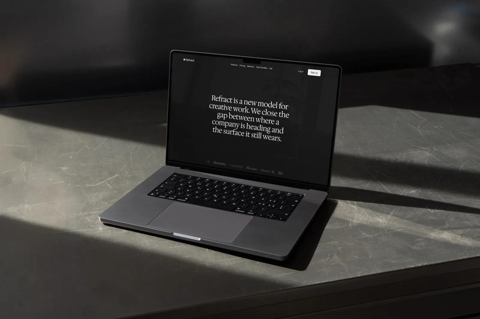 The laptop in low sun on a scratched metal bench, showing the all-black site with a serif statement set centred: "Refract is a new model for creative work."