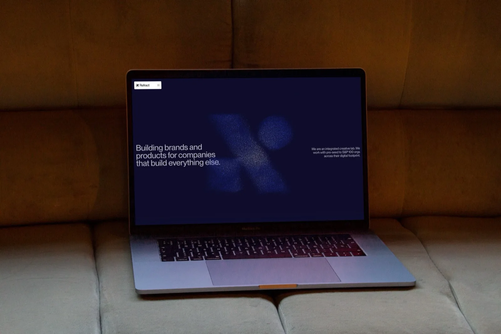 The laptop on a pale sofa showing the current site: the Refract mark held together out of blue particles on near-black, beside the line "Building brands and products for companies that build everything else."