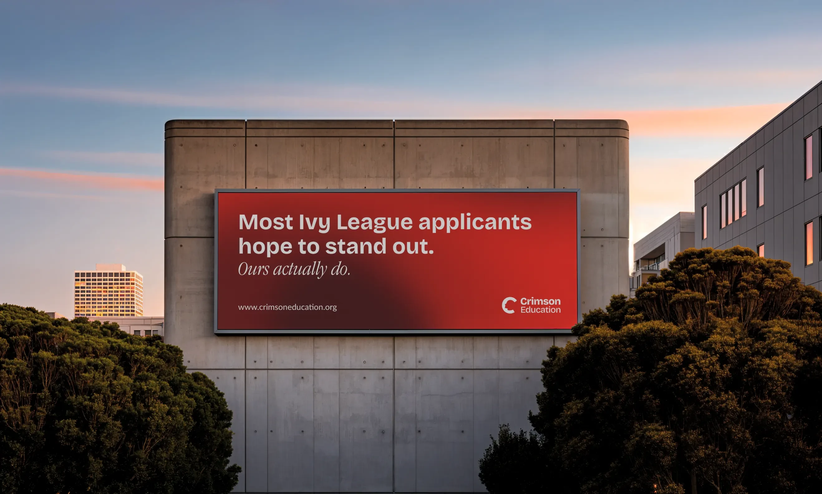 A lit billboard on a concrete facade at dusk reading “Most Ivy League applicants hope to stand out. Ours actually do.” in white on crimson