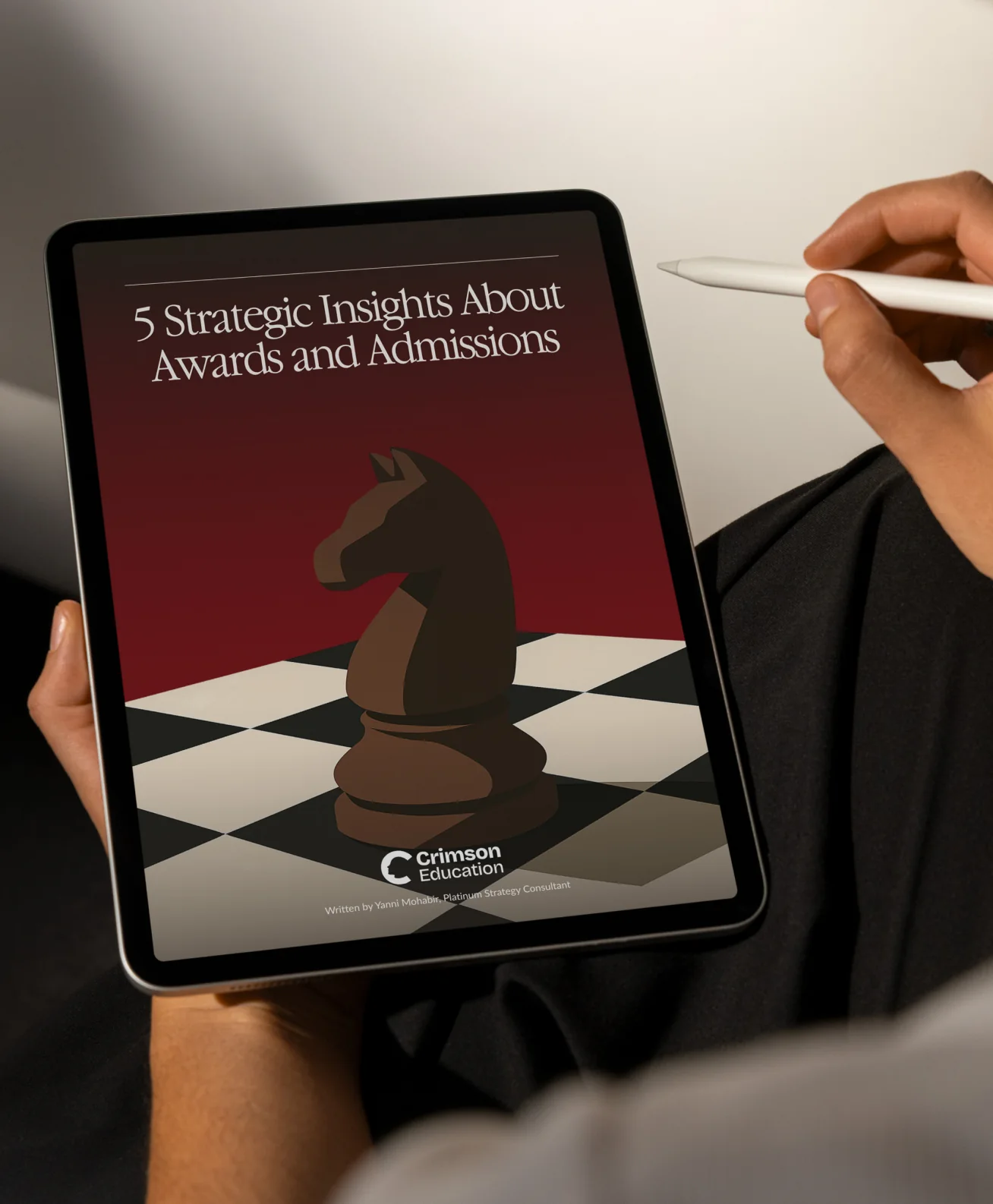 An iPad held with a stylus showing a Crimson ebook cover, “5 Strategic Insights About Awards and Admissions”, a chess knight standing on a board below the title