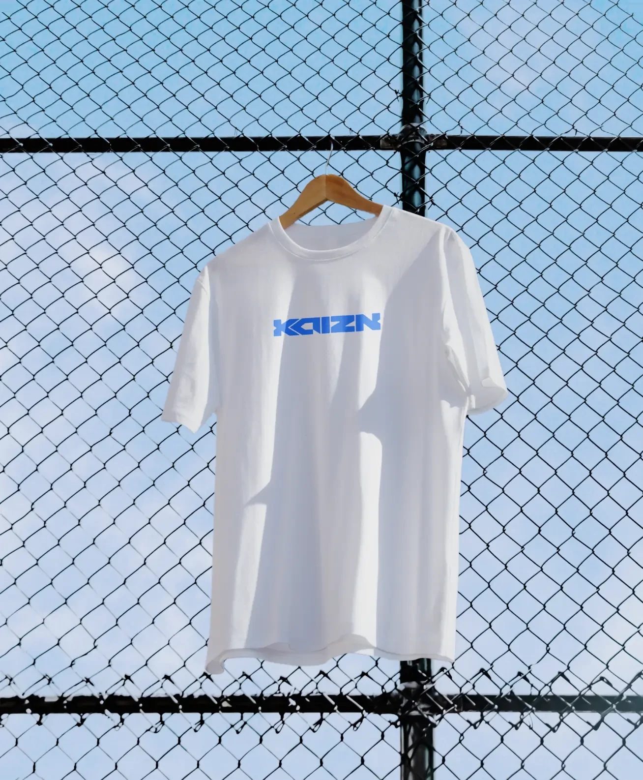 A white tee carrying the Kaizn wordmark, hung on a chain-link fence