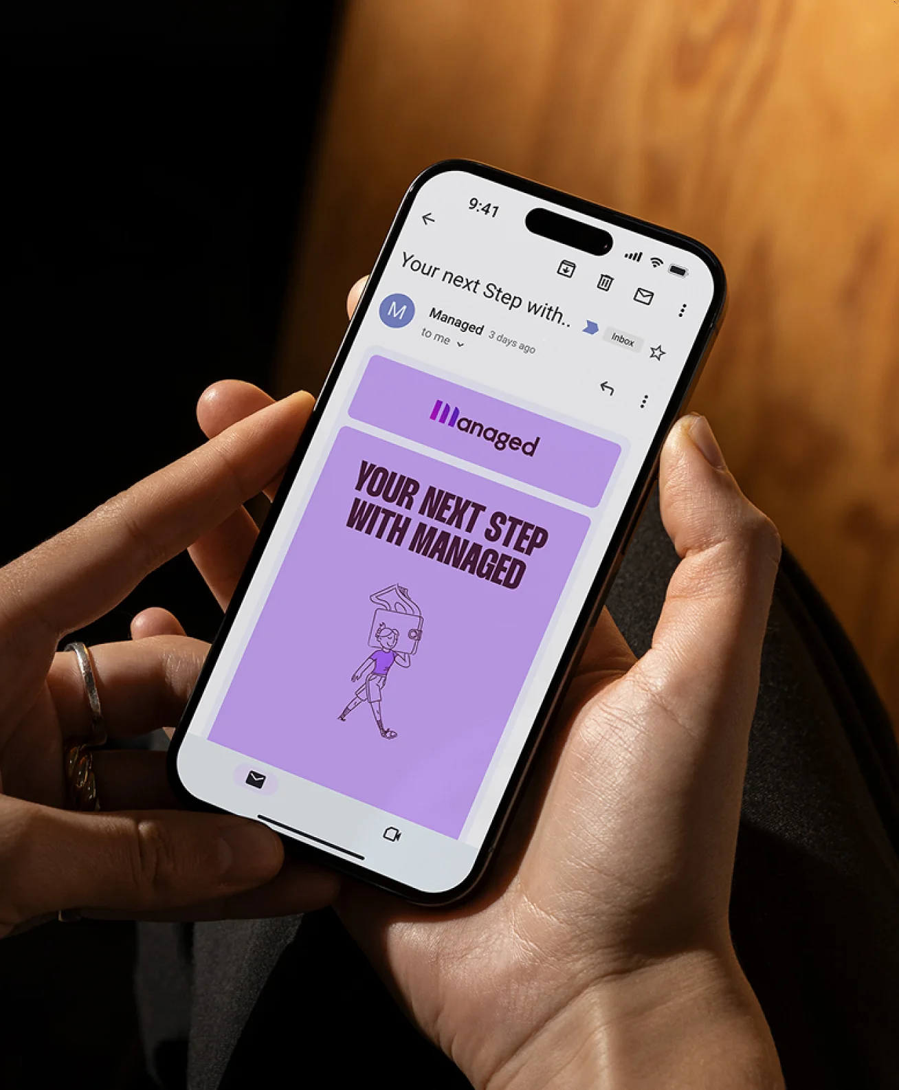 A phone held in two hands showing the invitation as it arrives: a lilac email card headed “Your next step with Managed” over a line drawing of a figure walking